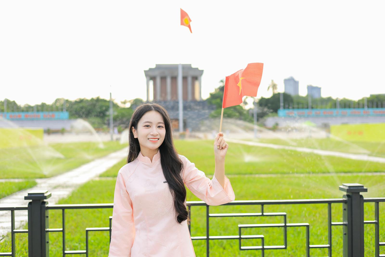 Vietnamese woman in traditional dress holding flag with Hanoi's landmarks in the background.