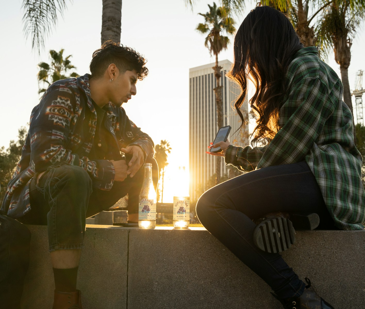 a man and a woman sitting on a wall looking at their cell phones