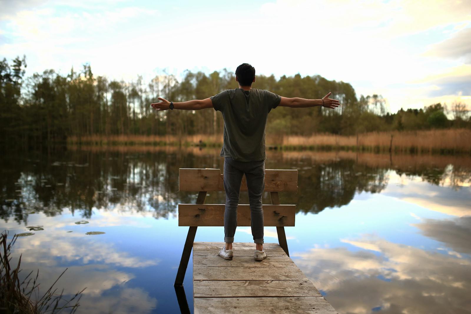 A person relaxes with open arms on a pier by a tranquil lake surrounded by nature.