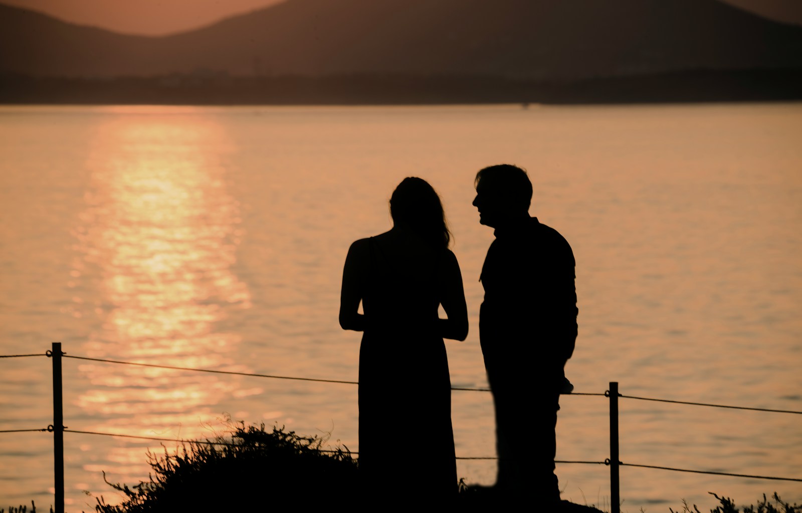 silhouette of man and woman standing near body of water during sunset
